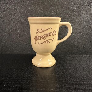 May include: A cream-colored Hershey's mug with a handle and a pedestal base. The Hershey's logo is printed in brown script on the front of the mug. The mug is made of ceramic and has a glossy finish.