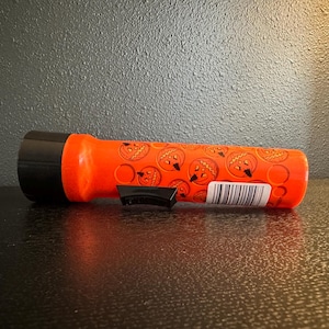 May include: Orange flashlight with a black head and a black switch. The flashlight is decorated with jack-o'-lantern faces. A barcode is visible on the side. The flashlight is lying on a dark surface.