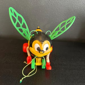 May include: A vintage pull-along toy bee with a black and yellow body, green wings, and red feet. The toy has a string attached for pulling and is made of plastic.
