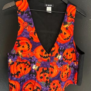 May include: A Halloween-themed vest with a V-neck design. The vest features a purple background with orange jack-o'-lanterns, bats, and small figures. The back and trim are black. The vest is buttoned and hanging on a black hanger.