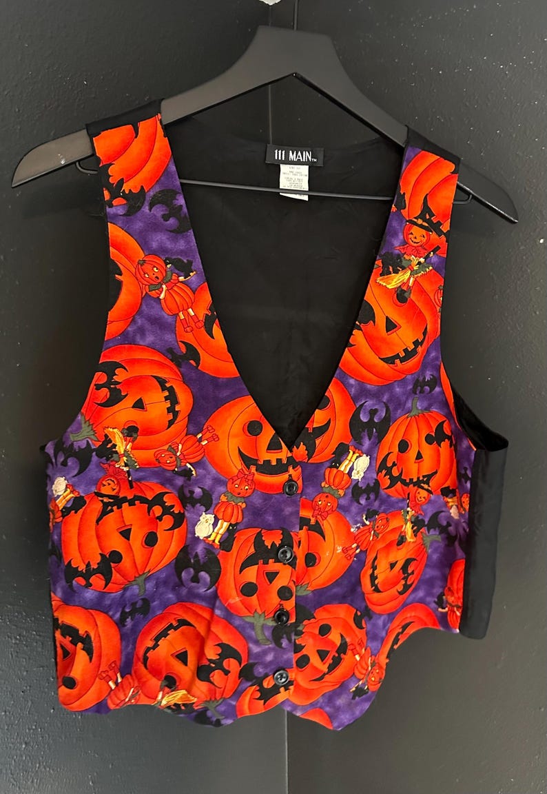 May include: A Halloween-themed vest with a V-neck design. The vest features a purple background with orange jack-o'-lanterns, bats, and small figures. The back and trim are black. The vest is buttoned and hanging on a black hanger.
