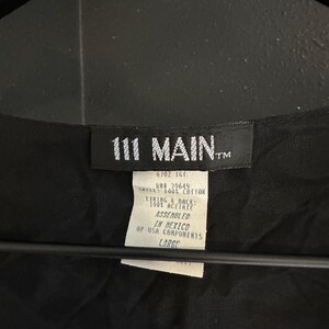 May include: Black garment tag with the text "111 MAIN" and a smaller tag with text including "SHELL: 100% COTTON" and "LARGE". The tags are attached to a black fabric, possibly a piece of clothing.