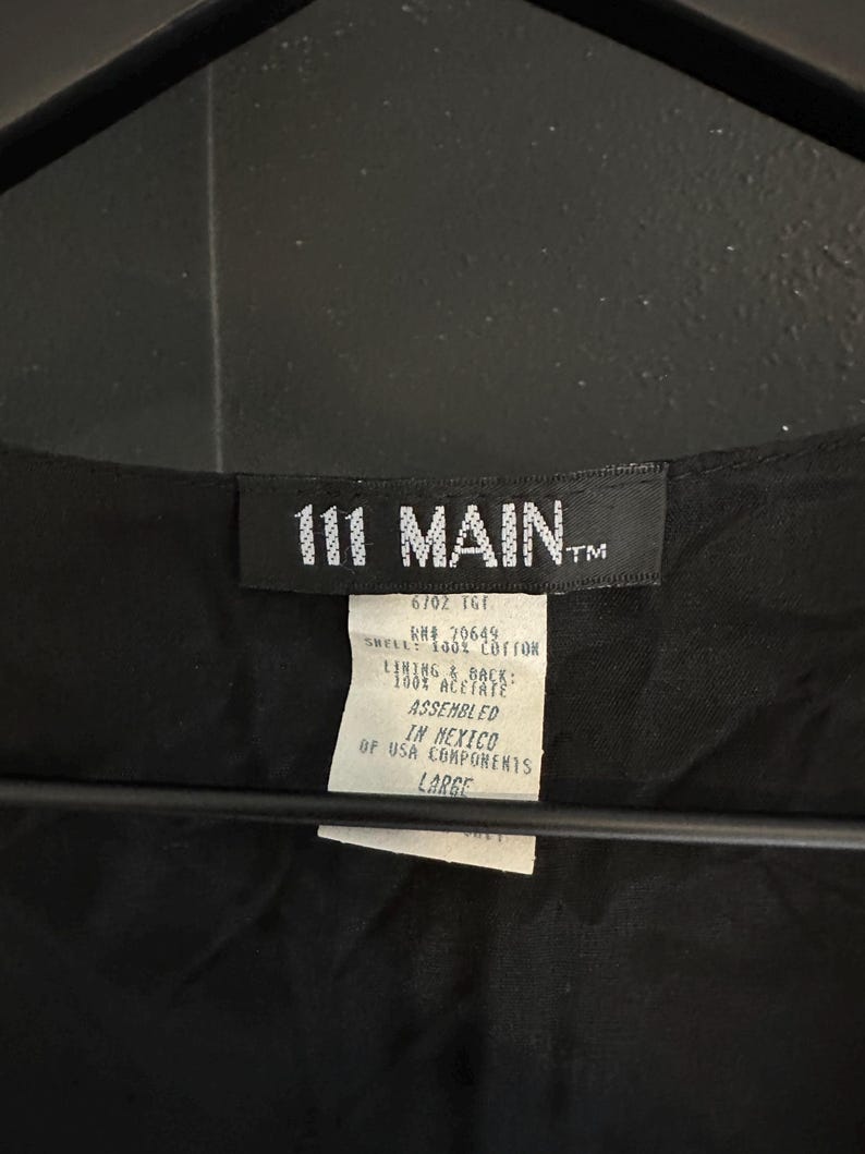 May include: Black garment tag with the text "111 MAIN" and a smaller tag with text including "SHELL: 100% COTTON" and "LARGE". The tags are attached to a black fabric, possibly a piece of clothing.