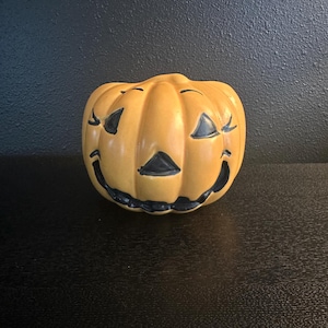 May include: A ceramic jack-o'-lantern with a smiling face. The pumpkin is painted a light orange with black accents for the eyes, nose, and mouth. The Halloween decoration is sitting on a black surface.
