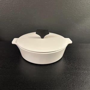 May include: A white, lidded casserole dish with a black handle. The dish has a rounded shape with small handles on either side. The lid is also white and fits snugly on the dish. The handle is a solid black color.