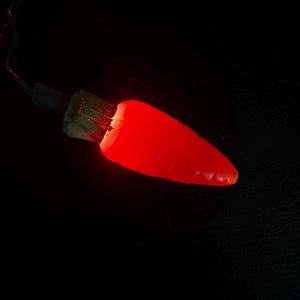 May include: A single red chili pepper-shaped Christmas light with a green base and a black wire. The light is illuminated, casting a bright red glow against a dark background.