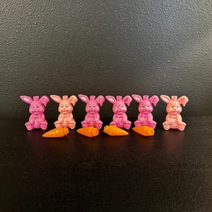 May include: Six small, pink bunny figurines with orange carrot accessories. The bunnies are in various shades of pink and peach, with upright ears and smiling faces. The carrots are bright orange.
