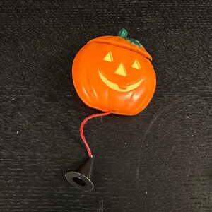 May include: Orange plastic jack-o'-lantern with a smiling face, triangular eyes, and a green stem. A red string connects the pumpkin to a black cone-shaped object. Halloween-themed decoration.