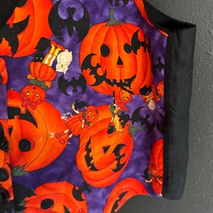 May include: A Halloween-themed vest with a purple background and orange jack-o'-lanterns. Black bats and small figures are scattered across the fabric. The vest has a black trim and buttons.