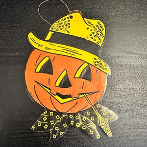 May include: A flat, die-cut cardboard wall decoration featuring a smiling orange jack-o'-lantern wearing a yellow hat with a black band and a black neckerchief with yellow diamond patterns. A thin gold string is attached to the top for hanging.