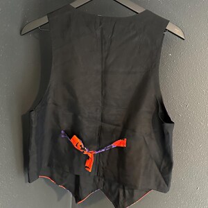 May include: Black vest with a red and purple patterned lining. The back of the vest is black, with an adjustable strap in red and purple. The vest is hanging on a black hanger.
