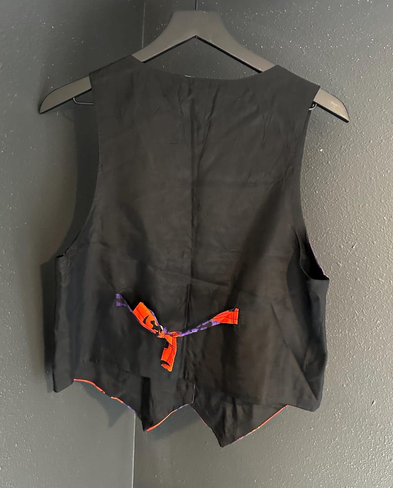 May include: Black vest with a red and purple patterned lining. The back of the vest is black, with an adjustable strap in red and purple. The vest is hanging on a black hanger.