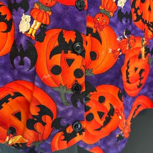 May include: A purple Halloween vest with an orange pumpkin pattern. The vest features black buttons and a repeating design of jack-o'-lanterns, bats, and pumpkin-headed figures. The fabric has a textured appearance.