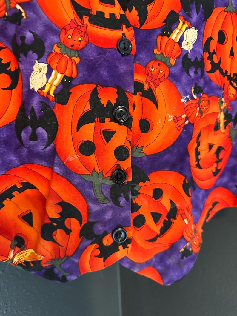 May include: A purple Halloween vest with an orange pumpkin pattern. The vest features black buttons and a repeating design of jack-o'-lanterns, bats, and pumpkin-headed figures. The fabric has a textured appearance.