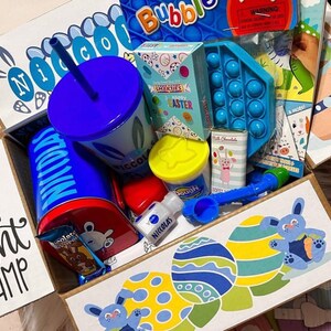 May include: A blue and white gift box filled with Easter-themed items, including a blue cup with a straw, a blue and yellow play dough container, a blue and white pop-it toy, and a blue and white Easter egg hunt champ sign.