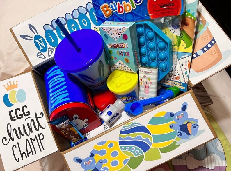 May include: A blue and white gift box filled with Easter-themed items, including a blue cup with a straw, a blue and yellow play dough container, a blue and white pop-it toy, and a blue and white Easter egg hunt champ sign.
