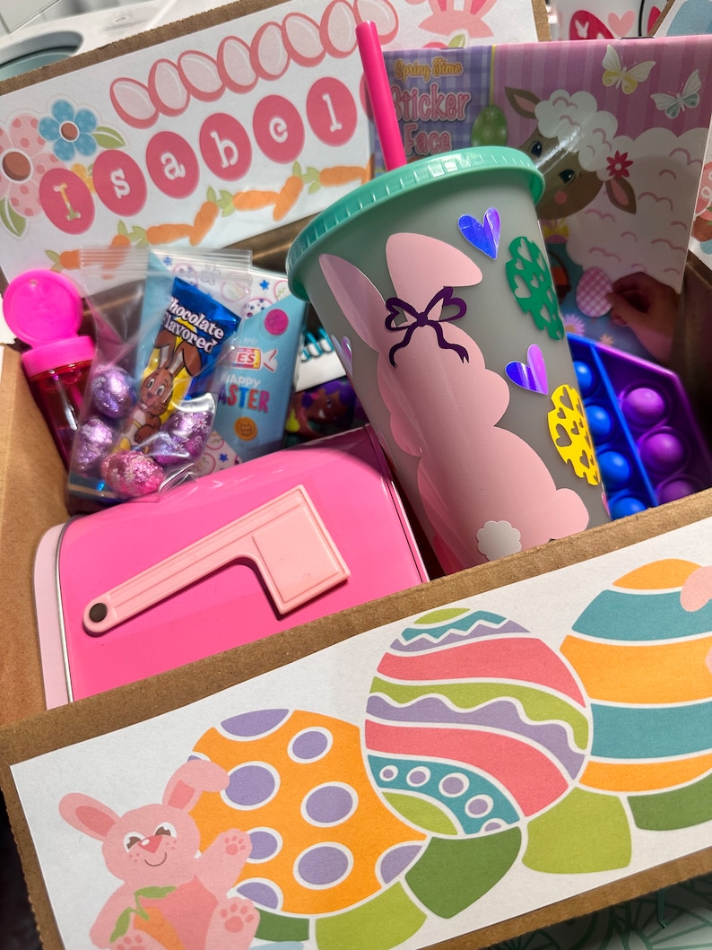 May include: A brown cardboard box filled with Easter-themed gifts. The box has a pink mailbox, a pink and green reusable cup with a bunny design, chocolate-covered candies, and a sticker book. The box is decorated with a sticker of a bunny holding a carrot and three colorful Easter eggs.