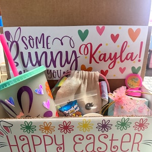 May include: A cardboard box with a white paper insert that says "Some Bunny Kayla!" and "Happy Easter" with colorful flowers. The box is filled with a variety of items, including a pink straw, a light blue cup with a bunny design, a small white bag, a chocolate bar, and a pink ribbon.