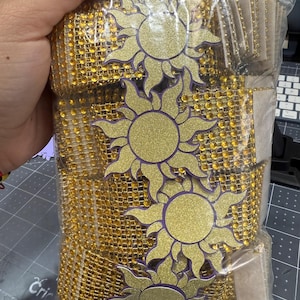 May include: A clear plastic bag filled with gold mesh ribbon and sun-shaped cutouts. The suns have a gold glitter center and purple outline. The ribbon is embellished with small, gold-colored rhinestones. The bag is held by a hand.
