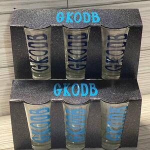 May include: Set of six clear glass shot glasses with the name "GKODB" printed in blue on each glass. The glasses are packaged in a black box with a silver glitter finish.