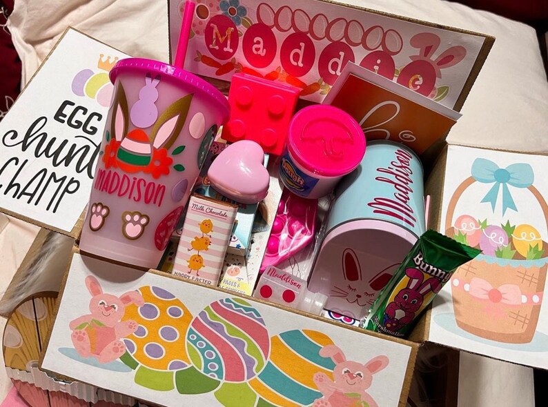 May include: A pink and blue Easter gift box with a personalized mug, a pink heart-shaped soap, a pink play dough container, a pink and white bunny, and a chocolate bar. The box also includes a green bunny-shaped candy bar, a pink and white bunny-shaped mug, and a pink and white bunny-shaped cup. The box is decorated with a pink and white bunny-shaped design and the words "Egg Hunt Champ" and "Maddison".