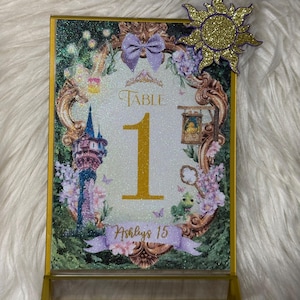 May include: A rectangular table number sign featuring a glittery finish, a gold frame, and a large gold number 1. The design includes a tower, a green lizard, a lantern, and a purple bow. The text reads Table 1 and Ashlys 15, with a gold sun cutout.