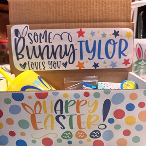 May include: A cardboard box with a yellow and white Happy Easter! design. The box contains a yellow cup with a blue star and the words "Some Bunny Tyler loves you" on a sticker.