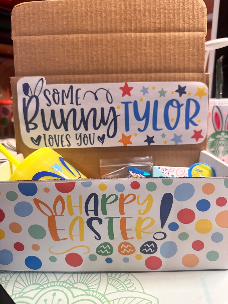 May include: A cardboard box with a yellow and white Happy Easter! design. The box contains a yellow cup with a blue star and the words "Some Bunny Tyler loves you" on a sticker.