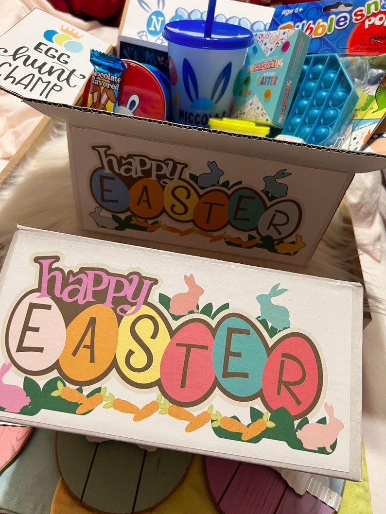 May include: A white gift box with a Happy Easter design featuring colorful eggs and bunnies. The box is filled with Easter-themed gifts, including a blue cup with a straw, a pop-it toy, and a chocolate-covered bunny.