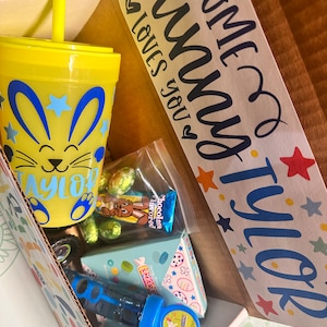 May include: A yellow and blue Easter-themed cup with a straw, a bag of chocolate-flavored candy, a blue bubble blower, and a box of candy with a bunny on it. The box is labeled "Some bunny loves you" and "Tyler".