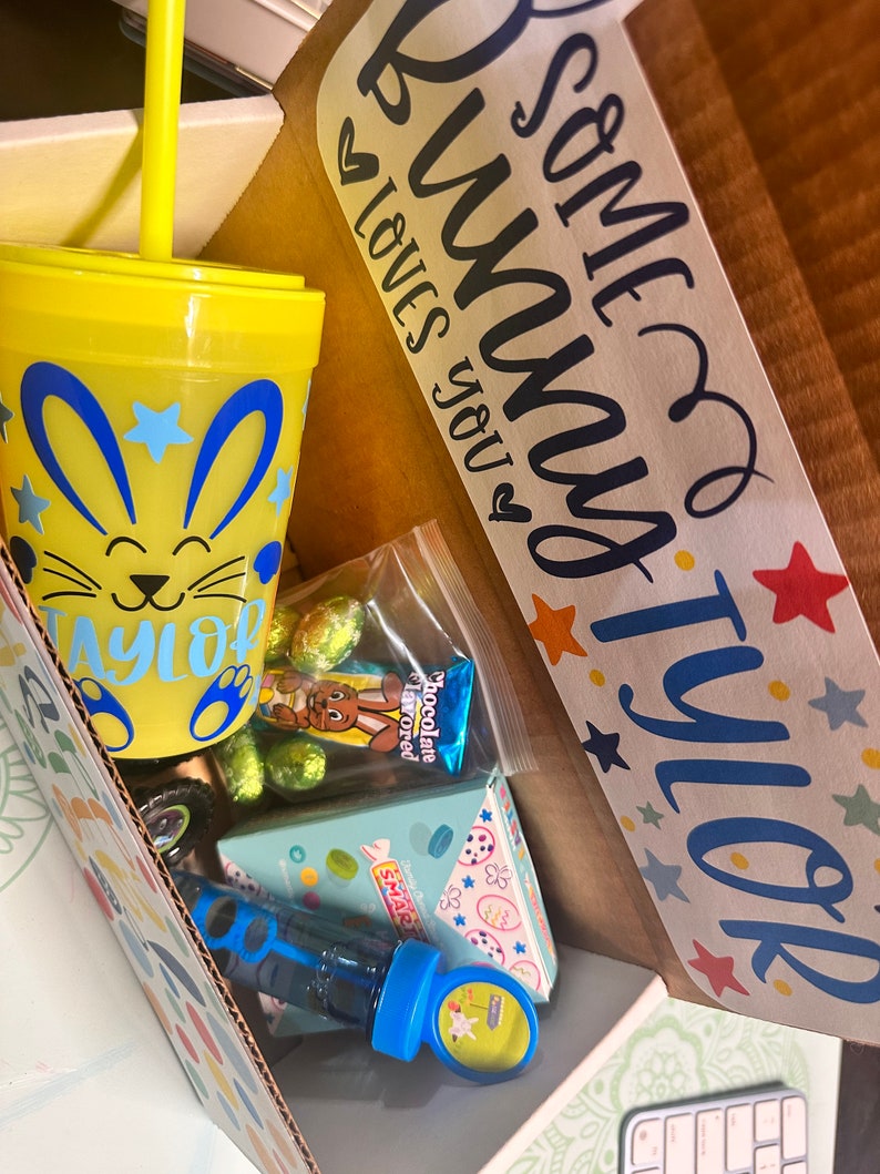 May include: A yellow and blue Easter-themed cup with a straw, a bag of chocolate-flavored candy, a blue bubble blower, and a box of candy with a bunny on it. The box is labeled "Some bunny loves you" and "Tyler".