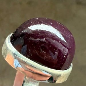 Op de afbeelding: Een zilveren ring met een grote, gepolijste, cabochon-geslepen edelsteen. De edelsteen heeft een diepe, rijke bordeauxrode kleur met een glad, afgerond oppervlak. De ring heeft een gepolijste zilveren band.