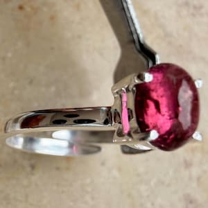 Puede incluir: Un anillo de tono plateado con una piedra preciosa redonda, pulida y de color rosa intenso, montada en un engaste de cuatro garras. La banda es lisa y metálica, con una superficie reflectante que capta la luz y resalta el color de la piedra.