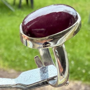Puede incluir: Un anillo de plata con una gran piedra preciosa de cabujón ovalada de color burdeos intenso, engastada en un bisel de metal pulido. La banda está hecha de un metal sólido de tono plateado reflectante, con un diseño limpio y acabado brillante.