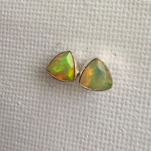 Könnte beinhalten: Ein Paar dreieckige Ohrstecker mit facettierten Opalen in silberfarbenem Metall. Jeder Opal zeigt ein Farbspiel mit grünen, gelben und orangefarbenen Lichtreflexen auf der Oberfläche der Steine, die in einer einfachen Metallfassung eingefasst sind.
