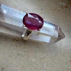 Könnte beinhalten: Ein Silberring mit einem großen, oval geschliffenen, tiefroten Edelstein in einer Vier-Krappen-Fassung. Der Ring liegt auf einer klaren, facettierten Quarzkristallspitze, die die leuchtende Farbe und die facettierte Oberfläche des Steins betont.