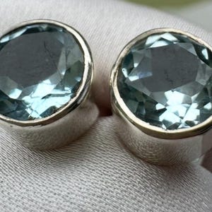 Peut inclure: Deux boucles d'oreilles à clous argentées, chacune ornée d'une grande pierre précieuse ovale, facettée, bleu clair. Les pierres sont serties dans une lunette en argent poli, créant un look classique et élégant. Les boucles sont présentées sur un tissu clair.