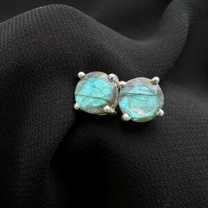 Puede incluir: Un par de pendientes de botón redondos con piedras de labradorita azul-verde iridiscente. Las piedras están engastadas en un metal plateado con cuatro garras. Los pendientes se muestran sobre un fondo de tela negra.
