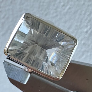 Puede incluir: Un anillo rectangular con una piedra preciosa transparente y una banda plateada. La piedra está facetada, creando múltiples superficies reflectantes. El anillo está sujeto por una herramienta de metal.