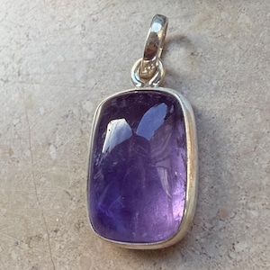 Può includere: Un ciondolo rettangolare in ametista viola con castone e anello argentati. La gemma lucida ha una forma arrotondata ed è incastonata in un design semplice ed elegante. Per collane.
