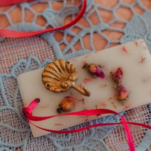 May include: A rectangular, off-white candle with dried rose buds and petals, and a gold seashell hook. A red ribbon is draped across a blue lace surface. The candle is a decorative item.