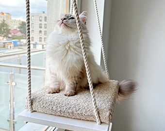 以下が含まれることがあります： 白い猫が窓から吊るされたベージュの猫用ベッドに座っています。猫用ベッドはロープ製で、柔らかくふわふわのクッションが付いています。猫はカメラの外にある何かを見上げています。