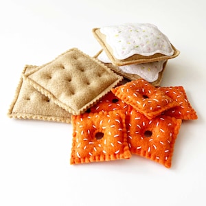 May include: Felt food play set featuring a stack of two graham cracker squares with white frosting and sprinkles, and five orange and white felt squares with a hole in the center.