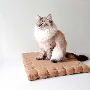 May include: A fluffy white and grey cat with blue eyes sits on a brown, quilted pet bed. The bed is square shaped and made of a soft, textured material.