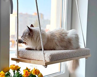 May include: A cat lounging on a hanging cat bed near a window. The bed is made of a light-colored wooden frame, a fluffy cream-colored cushion, and is suspended by rope. Yellow roses are in a vase in the foreground.