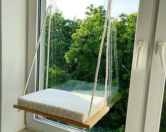 May include: A white cat bed hanging from a window using suction cups. The bed is made of a soft, white material and is suspended by white ropes. The bed is hanging in front of a window with a view of green trees.
