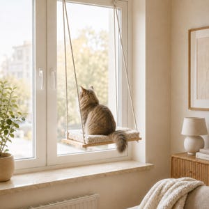May include: A rectangular wooden cat perch suspended by two thick ropes from the top of a window frame. The perch features a soft, beige, plush cushion on its surface, providing a comfortable resting spot for a long-haired brown tabby cat.