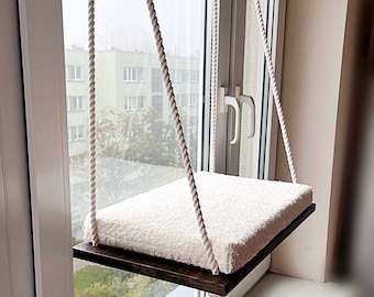May include: A white, fluffy cushion hangs from a window with white ropes. The cushion is attached to a dark brown wooden platform.