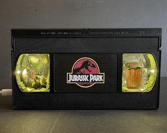 May include: A black VHS tape repurposed as a decorative light box featuring the Jurassic Park logo in the centre. The two side windows are illuminated, displaying miniature dioramas of a dinosaur in a jungle and the park entrance gates.