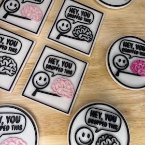 May include: White and black coasters with a smiley face holding a magnifying glass and a brain graphic. The text "HEY, YOU DROPPED THIS" is in a speech bubble. Some coasters have a pink brain, others a black brain.